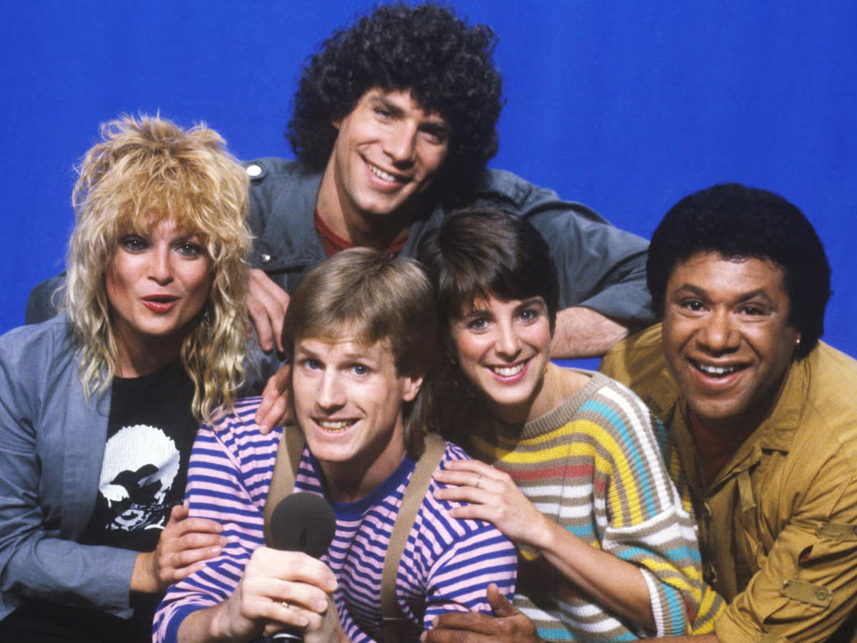 Ritratto dei cinque VJ originali di MTV degli anni '80: Nina Blackwood, Mark Goodman, Alan Hunter, Martha Quinn e J.J. Jackson in posa su sfondo blu.